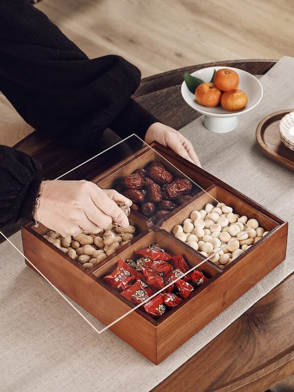 Wooden four-compartment snack box with clear lid filled with nuts and sweets on a dining table