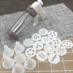 Cookie press piping gun with six nozzles and thirteen shaping discs laid out on a woven table mat