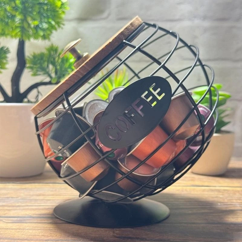 Tilted wire coffee capsule holder with wooden lid filled with capsules on a wooden table beside plants